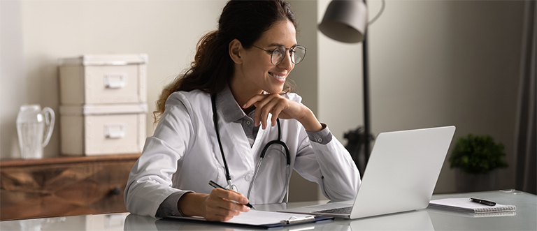 Smiling professional female doctor taking notes, looking at laptop screen Smiling professional female doctor wearing glasses and uniform taking notes in medical journal, filling documents, patient illness history, looking at laptop screen, student watching webinar