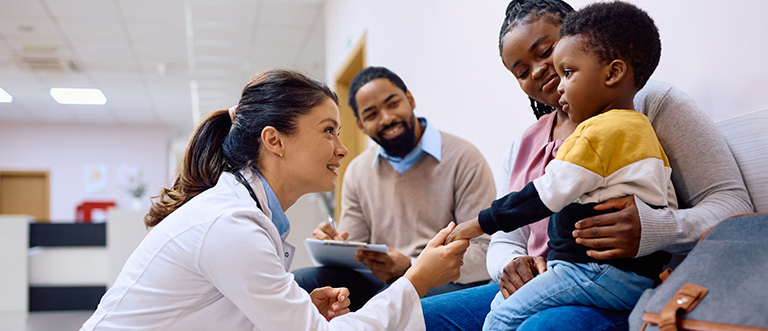 Female pediatrician talking to black little boy who is sitting on mother's lap at the clinic. Smiling doctor holding hand on African American boy who came with parents for medical exam at pediatric clinic.